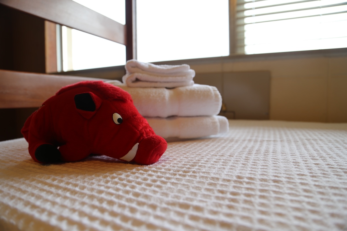 A razorback plush sits on a clean white bed that was made by a conference ambassador.